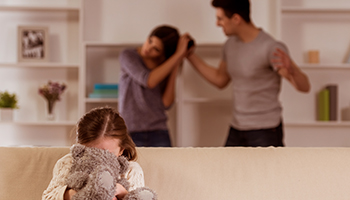 a young girl holds her teddy bear tightly, her expression anxious as a couple has an argument